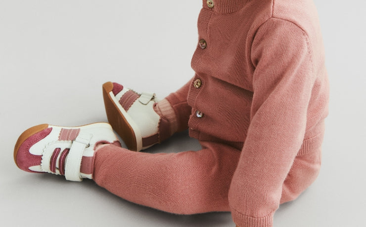 Child wearing a pink knitted outfit with white and red shoes on a light gray background
